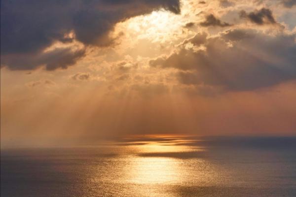 Vast, calm Ocean as far as the eye can see under dark clouds with rays of orange sunlight shining down.