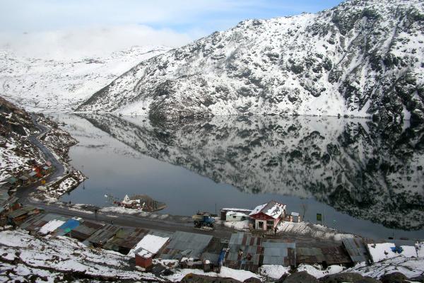 A lake surrounded by snow covered mountains with tin roof huts and a road along its perimeter in the foreground under blue skies and white clouds..