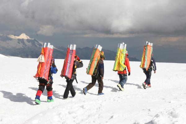 Five people, each carrying a group of long cylindrical tubes on their backs, walking away in a line on snow covered ground with snow capped mountain range at a difference under gray cloudy sky.