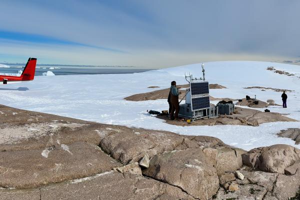 A small plane in a snow-covered ground with the background of sea with icebergs in it and people and a bedrock in the foreground working on equipment.