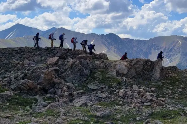 A group of eight with backpacks and walking gear walking along a rocky path with a backdrop of mountains, under blue skies and white clouds.
