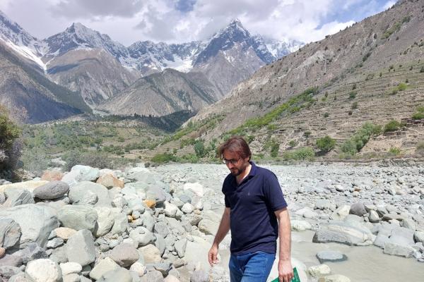 A man holding a green-covered notebook walking in a rocky river bed with a little stream flowing behind him and sparse tree covered hills and mountains in the background. 