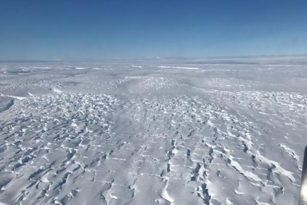 Vast area covered in snow and ice under blue skies with a jet engine visible in the bottom right corner. 