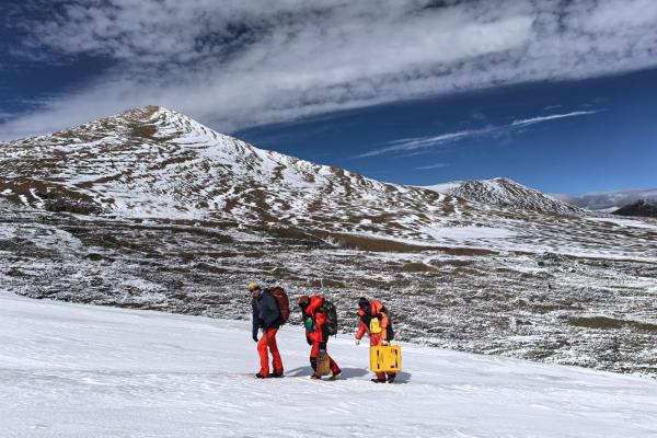 Three people walking up a snow covered hill, surrounded by mountains, with blue skies and white clouds..