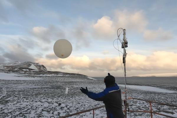 A person releasing a weather balloon into the sky from a snowy landscape during sunrise or sunset.