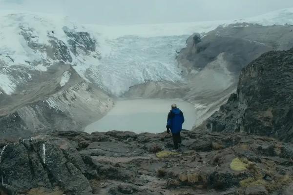 Person in blue jacket standing on rocky ground, observing a glacier and a glacial lake in a vast, mountainous landscape.