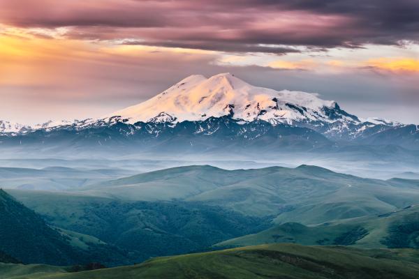 view of Elbrus mount in the morning at sunrise with a dramatic cloudy sky and foggy green highland meadows. Summer mountain landscape. North Caucasus, Karachay-Cherkess Republic, Russia