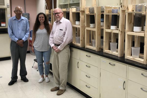 Three individuals standing in a laboratory with shelves containing scientific samples behind them.