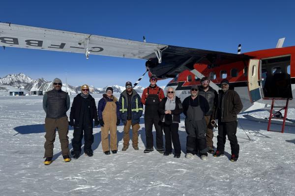 Group of ten people standing in front of a red and white plane on a snowy landscape with mountains in the background.
