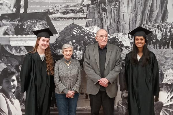 Two people in caps and gowns flank two adults in front of a photo montage.