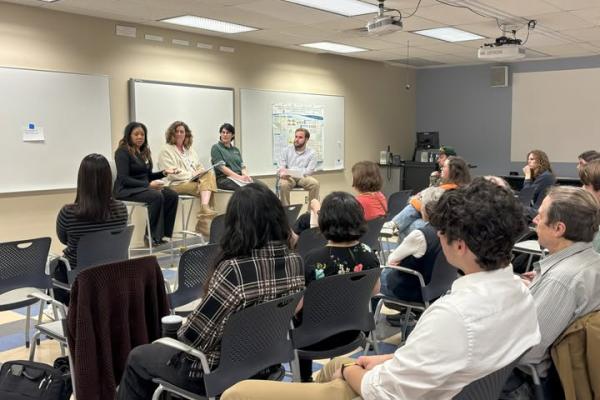 Four panelists are seated in front of a classroom, engaging with an attentive audience during a discussion.
