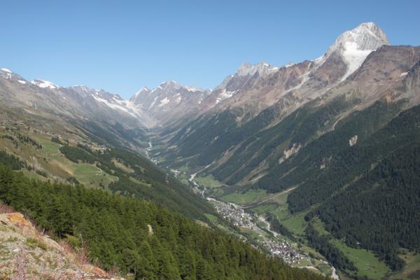 A wide valley with alpine villages in the middle surrounded by steep forested slopes and high mountain peaks with glaciers.