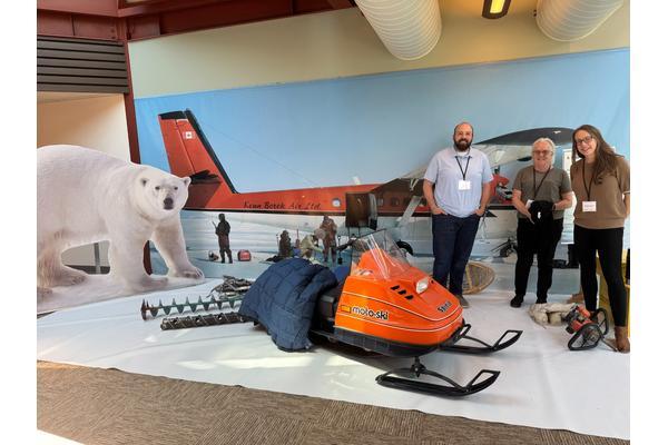 Three people stand smiling in front of a polar fieldwork display.