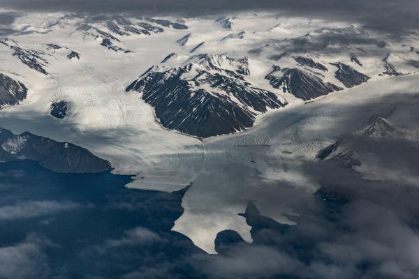 Here’s a short version for social media accessibility:  **Short alt text:** Glacier flowing between dark mountains into the ocean under clouds.