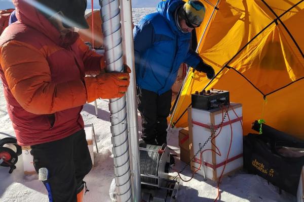 Two people are setting up a tall drill Ice core beside an orange dome tent.