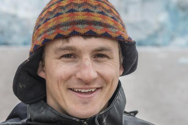 Close-up of Alex Michaud smilingwearing a colorful knit hat and a black jacket, with a blurred icy outdoor background.
