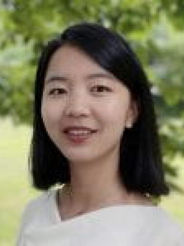 Yanlan Liu with white blouse smiling with blurred green tree, shrub background 