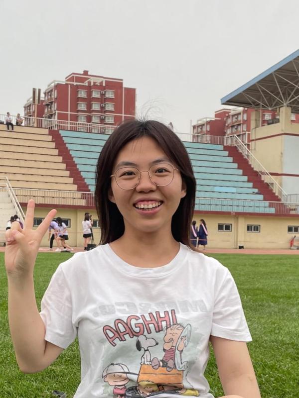 Yue Zhang smiling with glasses and white graphic tee holding piece sign in sports field grass