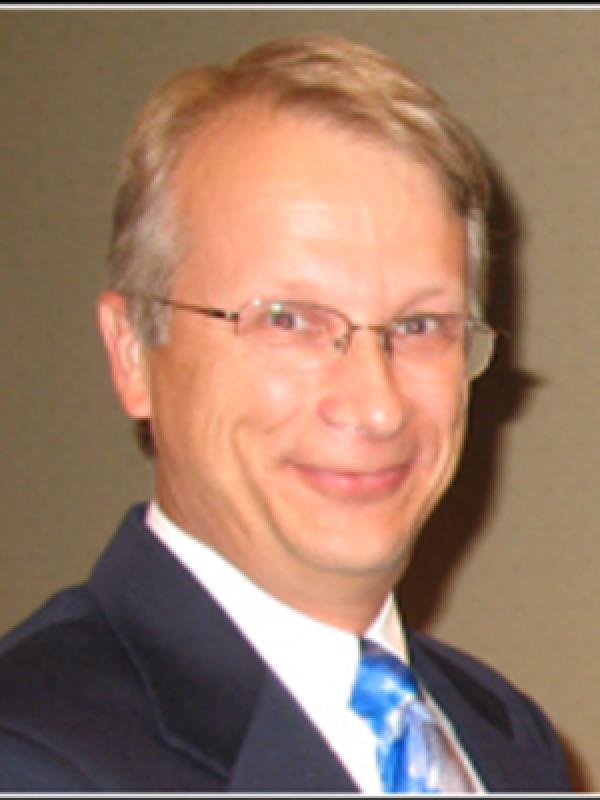 Left side view above chest portrait of John Olesik smiling with short blonde hair wearing glasses and a dark gray  suit and white collar shirt and blue/brown multi color tie