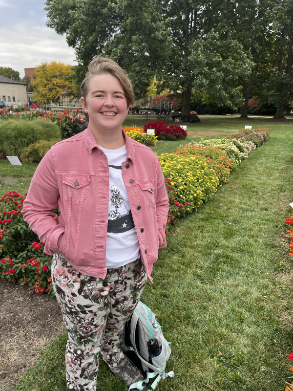 Savannah, a young person in a pink jacket and floral pants, standining in a garden with flowers and trees behind her