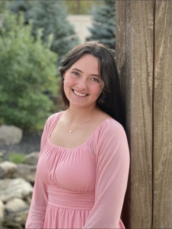 Hannah Fox wearing a pink dress smiling next to a tree with more trees in the background. 