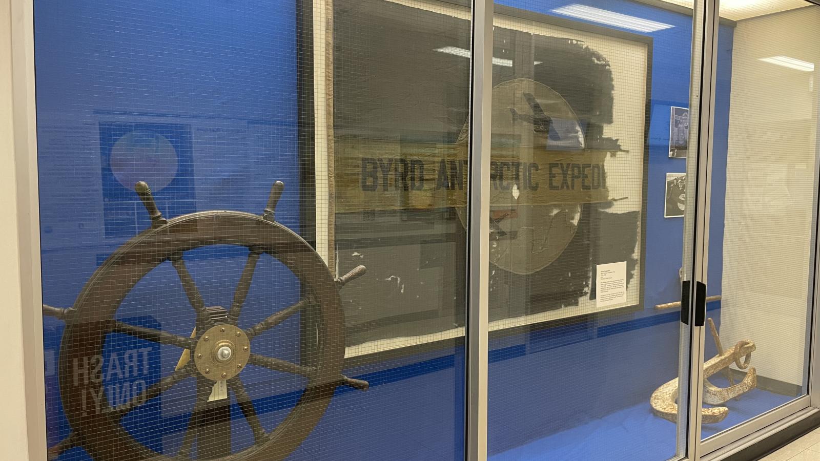 A side view from left of a full length built in display case outside of the Goldthwait Polar Library including a ship's large white anchor, a ship's steering wheel in dark wood and a large framed fabric shredded flag and 2 side images on a blue background behind a glass display