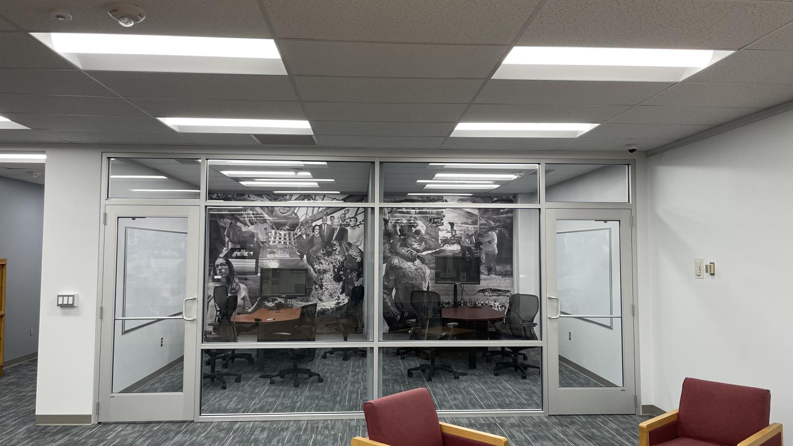 image taken from inside the Goldthwait Polar Library in the far back 2 glass enclosed quiet rooms  with a wall length vinyl drape of images on the back wall with some chairs and desk inside and gray carpet, white walls and a light wood table and 2 lounge chairs with red fabric in foreground on bottom right 