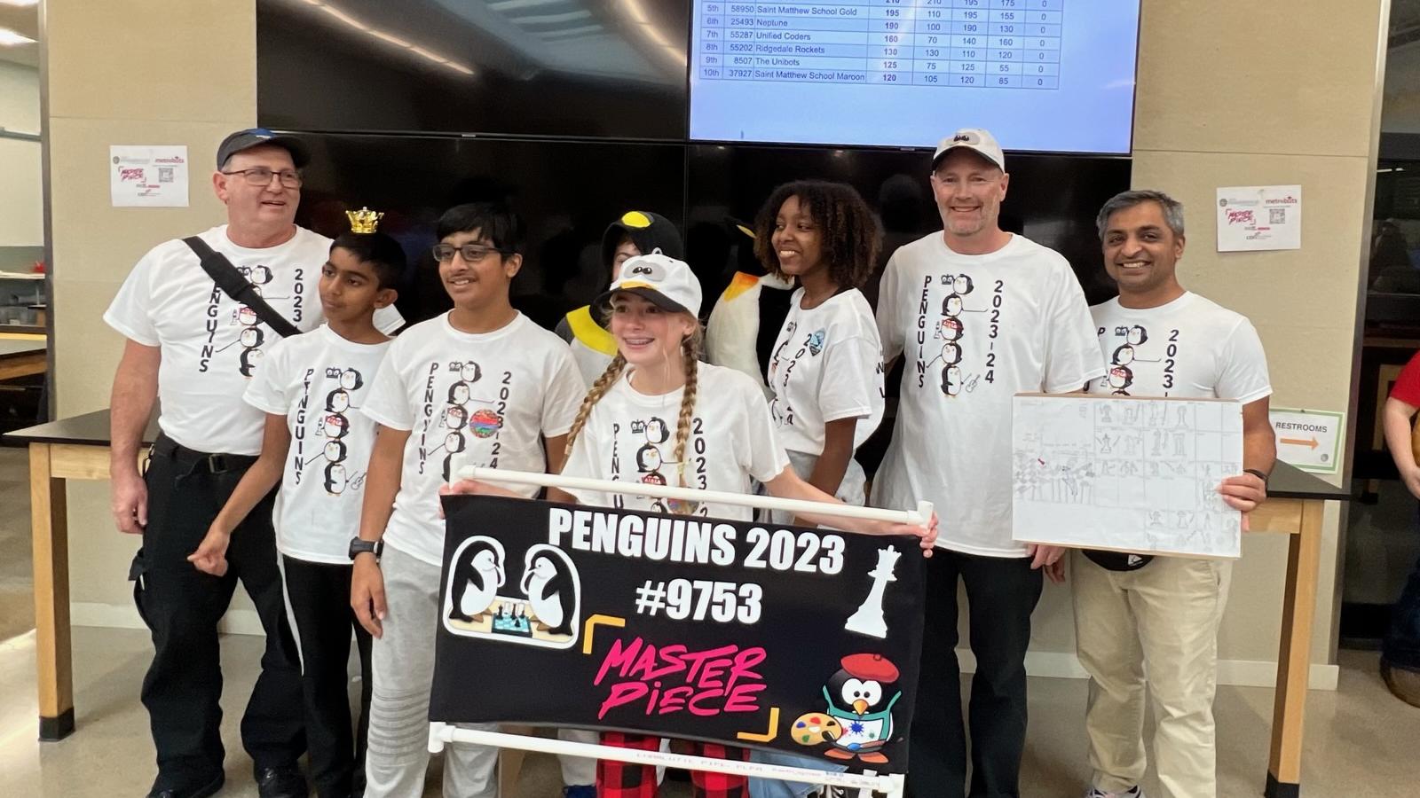 A group of youth and adults posing indoor for a picture with one youth holding an banner with images of penguins and a chess piece and an adult holding a plaque,