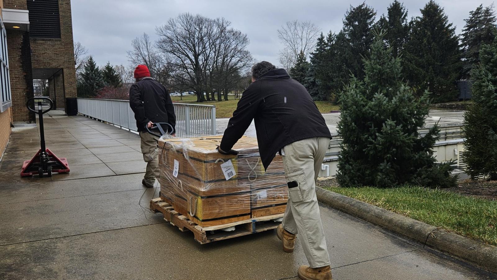 Two men rolling a cart with a pallet of wooden boxes along a sidewalk outside.