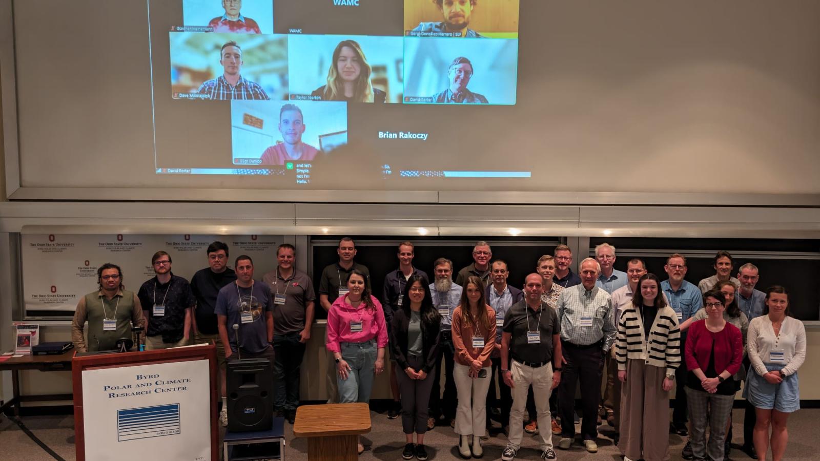 Group photo of a crowd in a lecture hall, standing on stage with a back ground of people in Zoom sessions on the big screen.