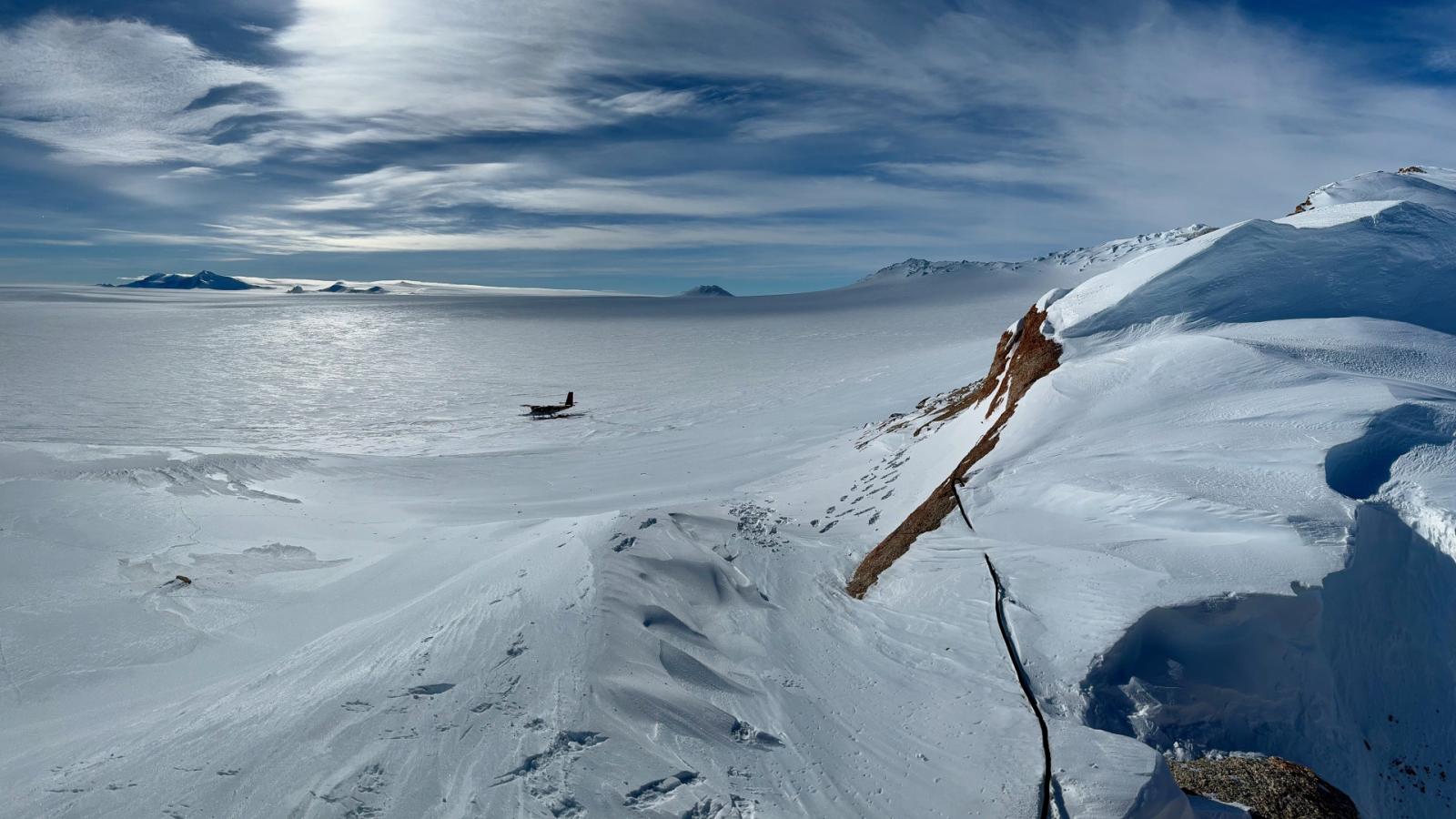 A vast area covered in snow and ice with mountains and a small plane in the snow at a distance.