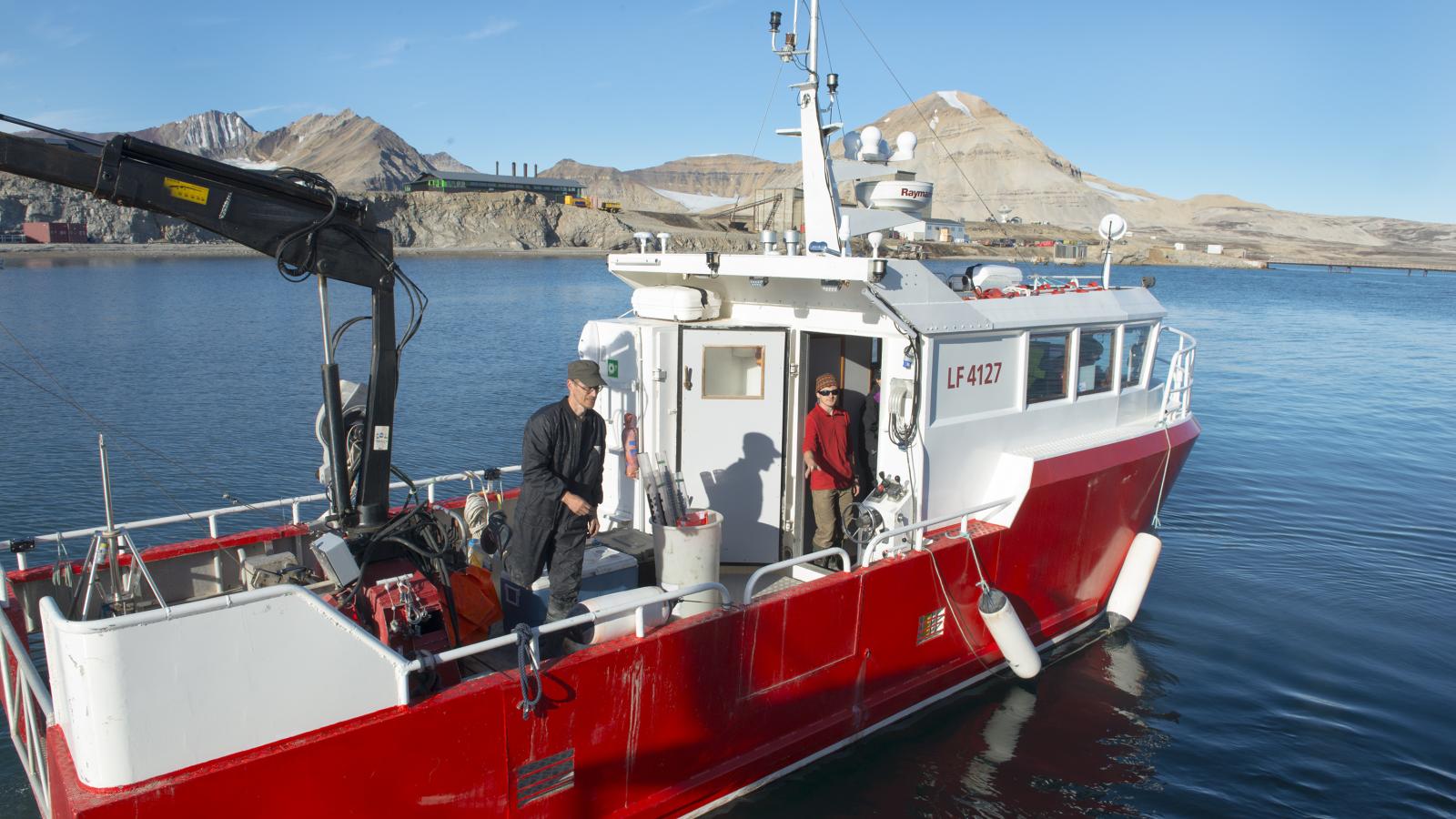 A small red and white boat on blue waters with rocky mountains and some industrial buildings in the background under blue skies