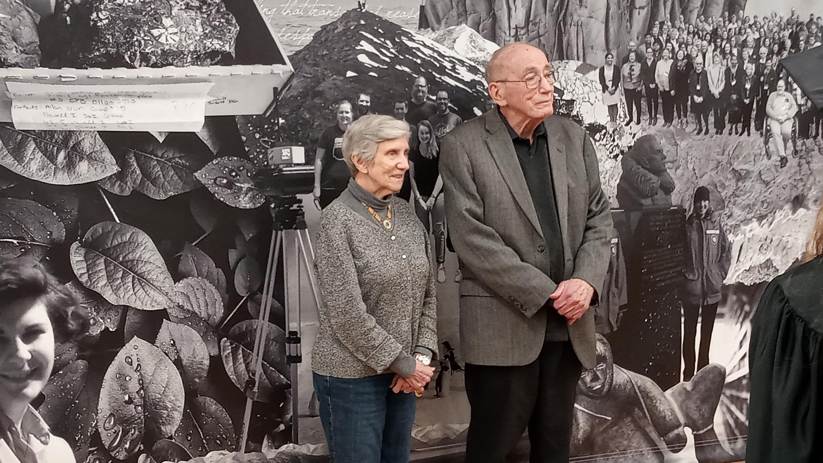 Two individuals standing in front of a large black and white photo collage featuring historical images and photographs of people.