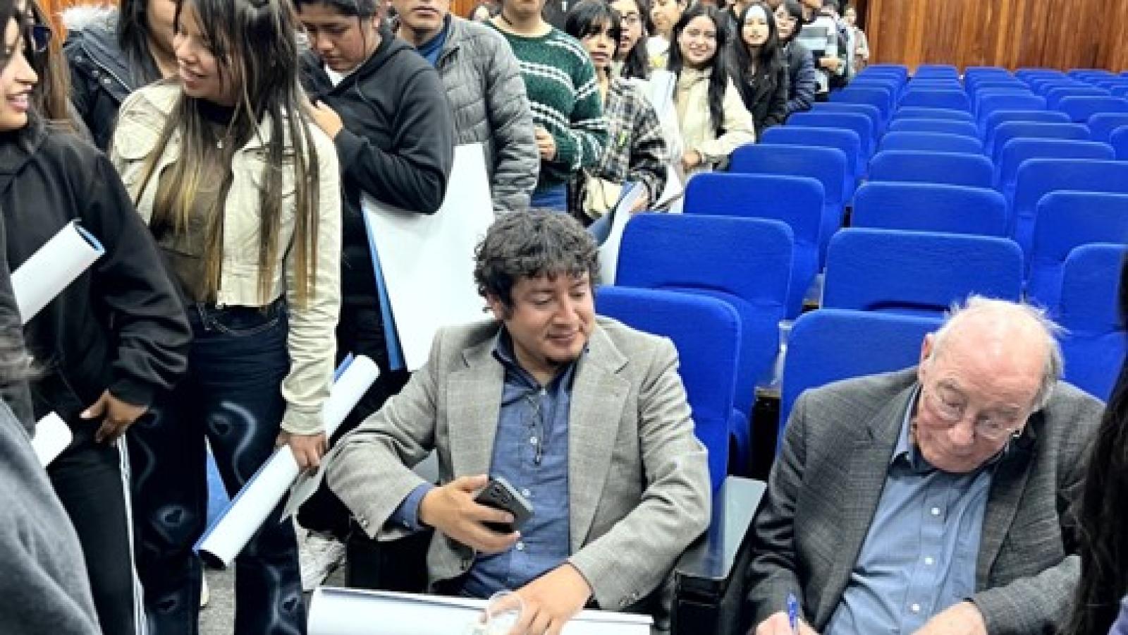 Students line up in a lecture hall as two speakers seated in blue chairs sign posters and papers.