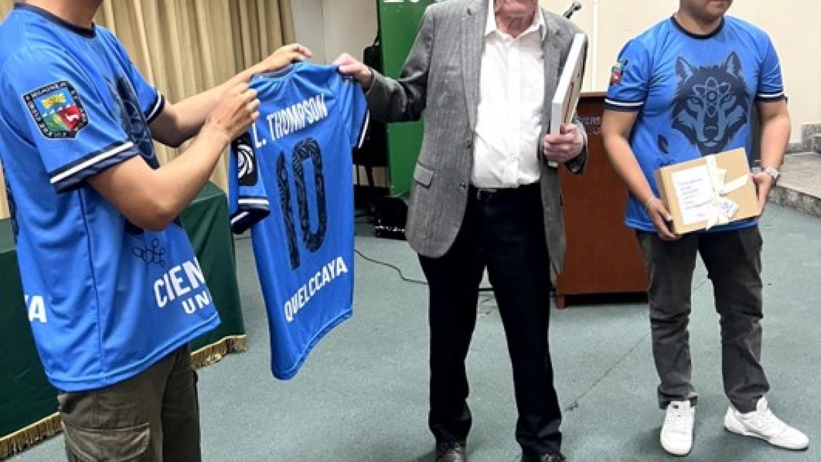 Two students in blue jerseys present a personalized soccer jersey and gift to an older guest in a gray blazer at the front of an auditorium.