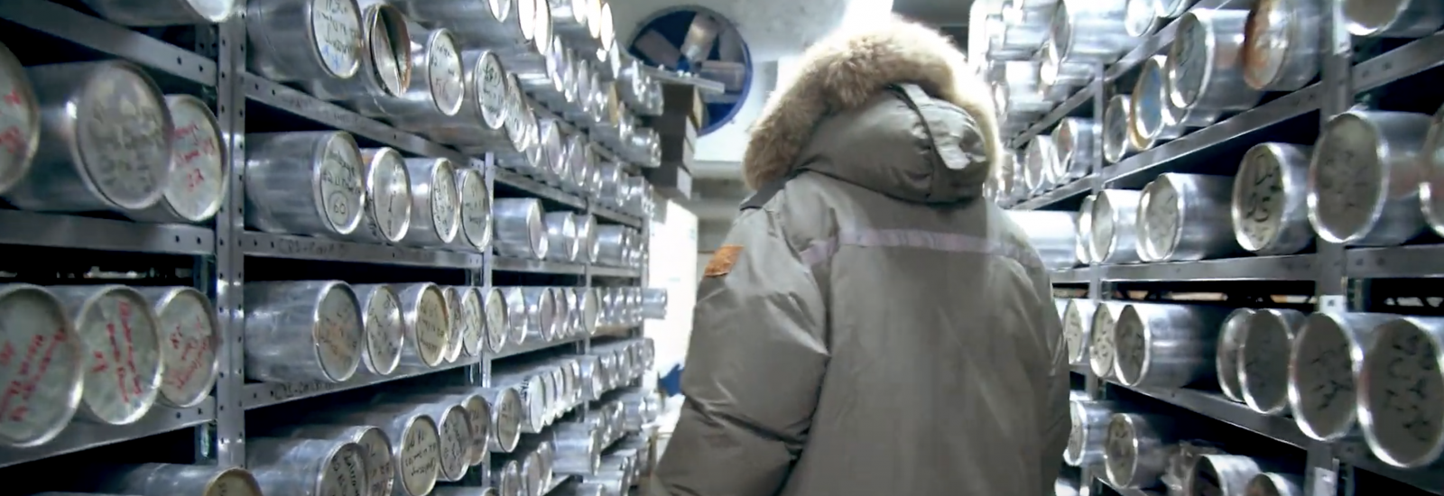 Lonnie Thompson from behind wearing a grey winter coat. He is walking through the ice core freezer and is surrounded by ice cores (long silver cores) on shelves on both sides.