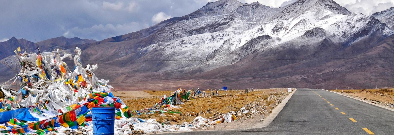 Blue trash can on paved road with colorful fabric trash pieces all over and snow covered mountains at a distance