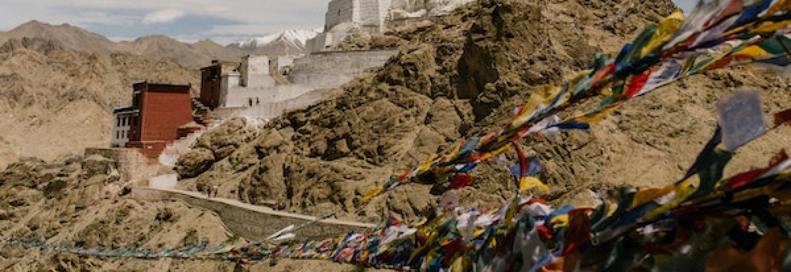 prayer flags of different sizes and colors on ropes flow in the wind fixed on both sides dominating the right side of the image with a series of white buildings cascading down the mountain or rock and dirt and a brown building at the bottom with blue sky and white  feather clouds