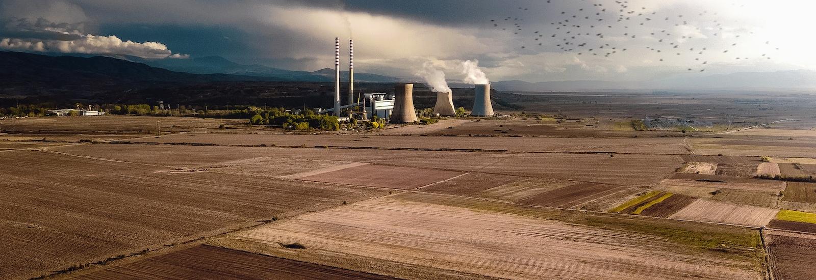 landscape image of bare farmland with brown shades of earth and a nuclear power plant with 3 stacks at a distance with mountains and dark and light clouds and sky with a flock of birds flying and a tree line and buildings around the plant