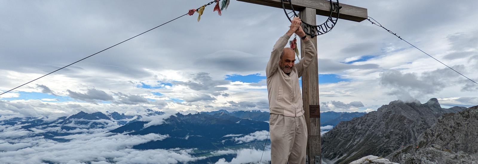 Henry Brecher, an old man in beige clothes, standing on the edge of a mountain with his hands clasped above his head victoriously