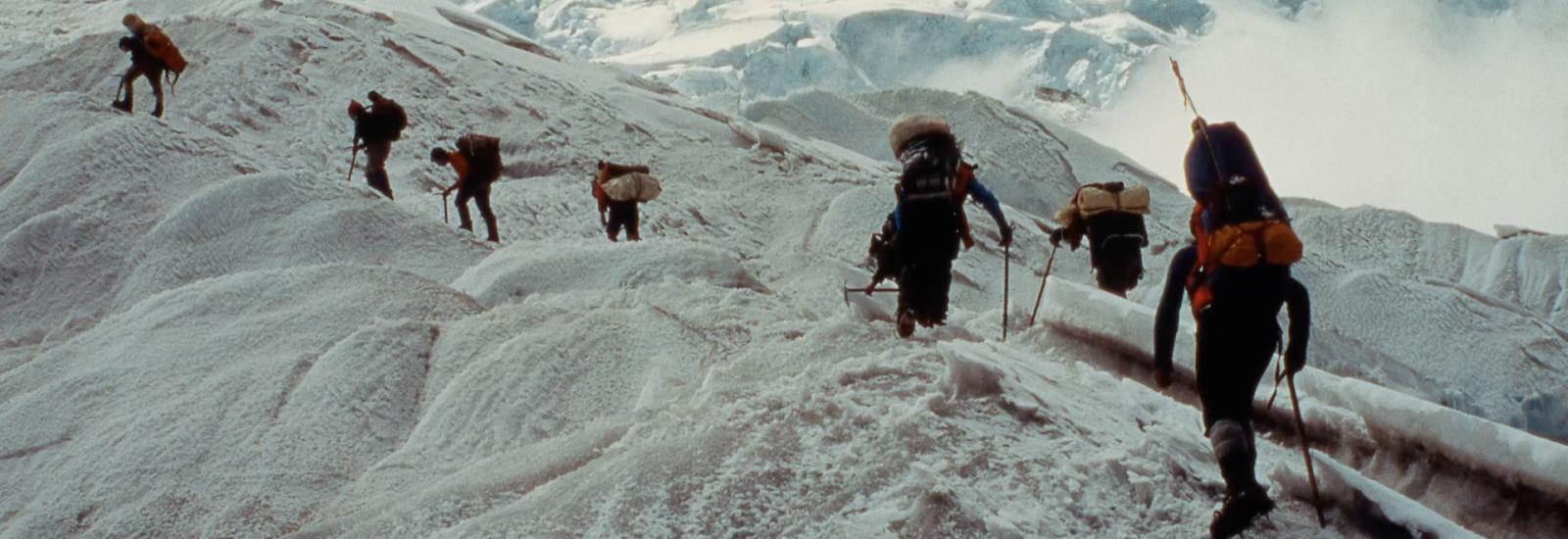 People walking up a mountain on ice carrying climbing gear surrounded by snow and mountains.