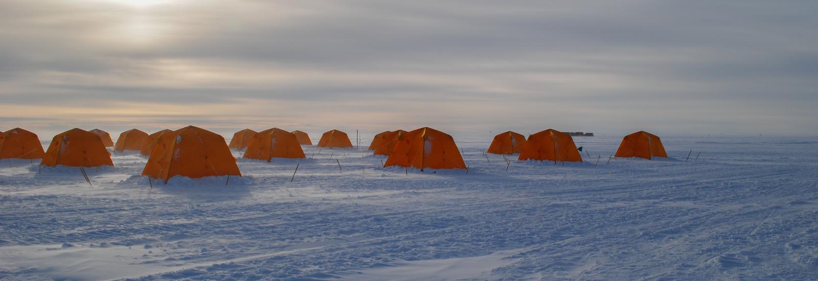 Snowy field with a group of orange tents under cloudy skies with halo of sun.
