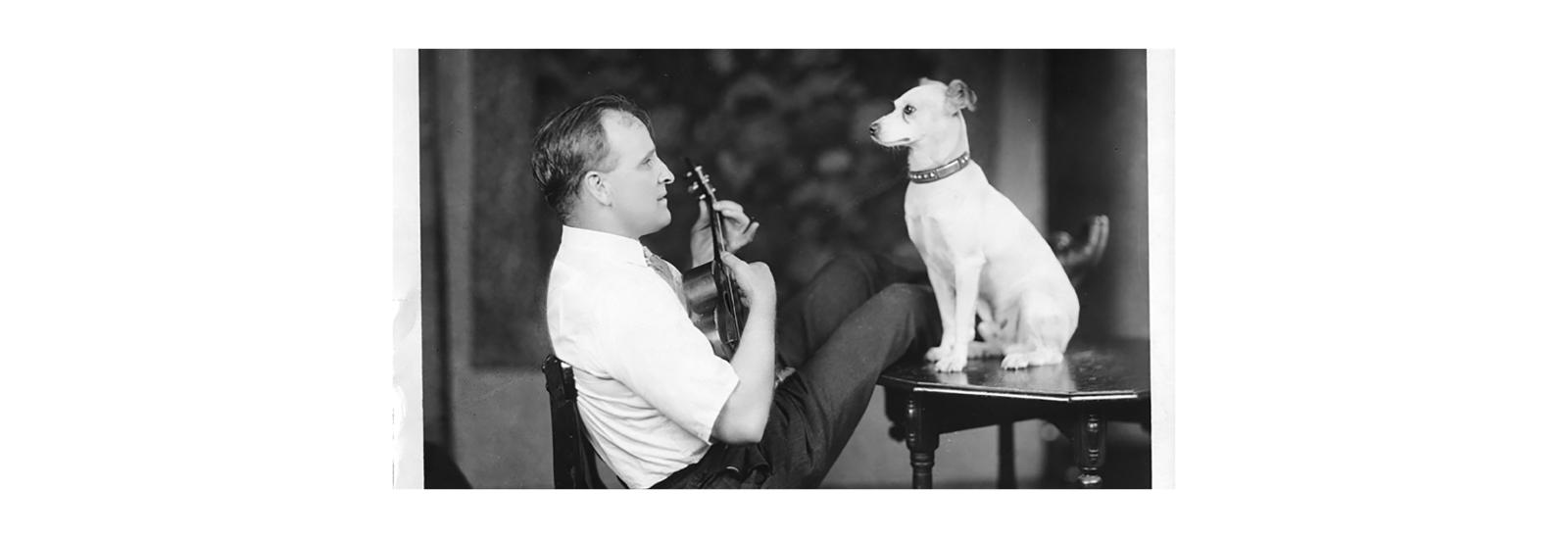 A man sitting at a table with his legs stretched on the table, playing a ukulele with a dog facing him sitting on the table in front of him.