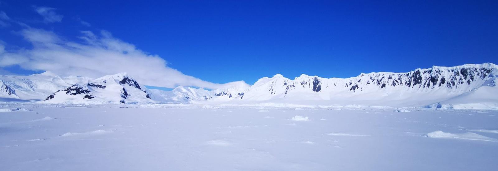Snow covered ground and mountains in a distance under blue skies and white clouds.