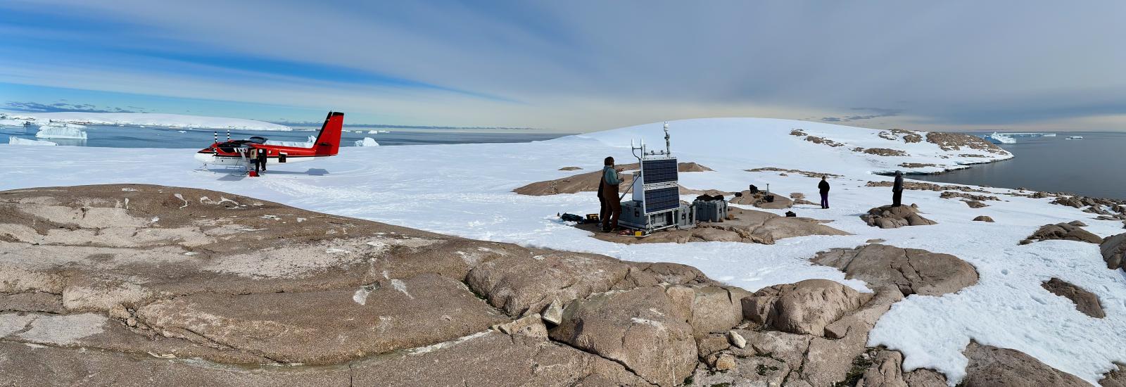 A small plane in a snow-covered ground with the background of sea with icebergs in it and people and a bedrock in the foreground working on equipment.