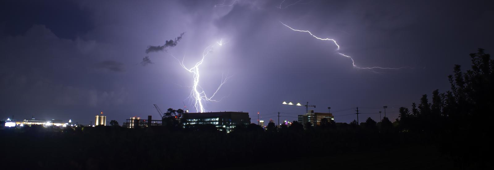 Bolts of lightning lighting up the night sky in the distance as a backdrop to taller buildings among tree lines.