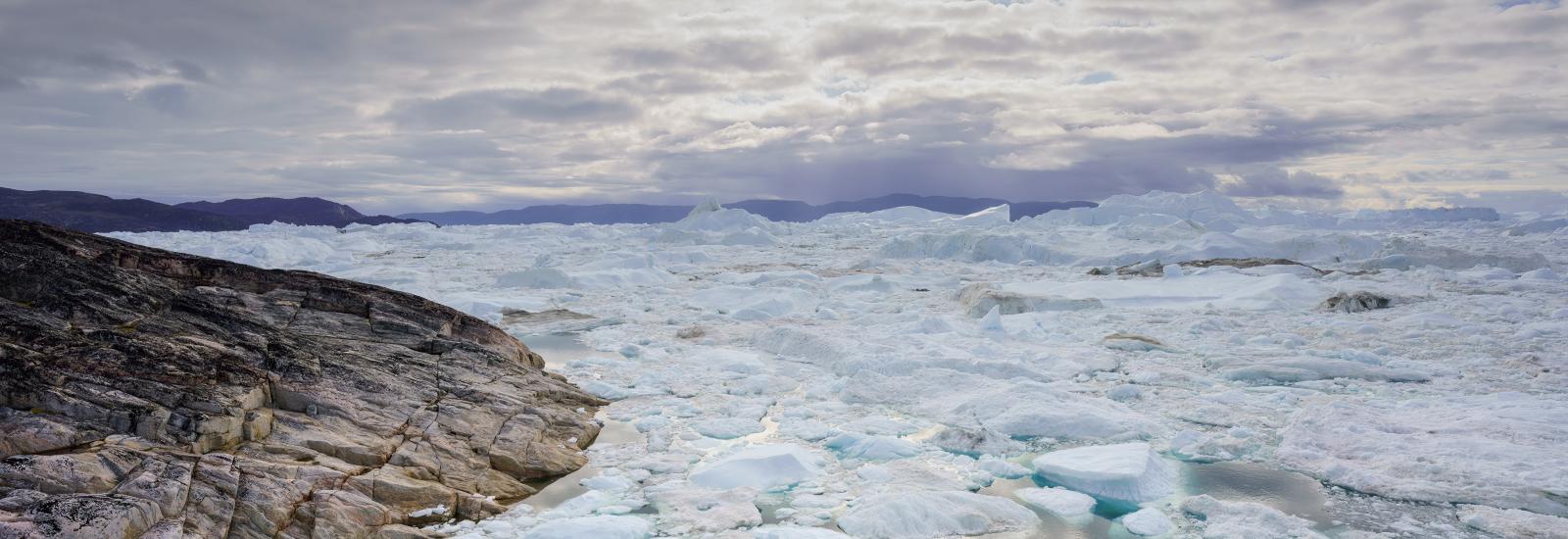 An Icefjord in Greenland, with large chunks of ice floating on the water under a cloudy sky, with rugged terrain in the foreground.