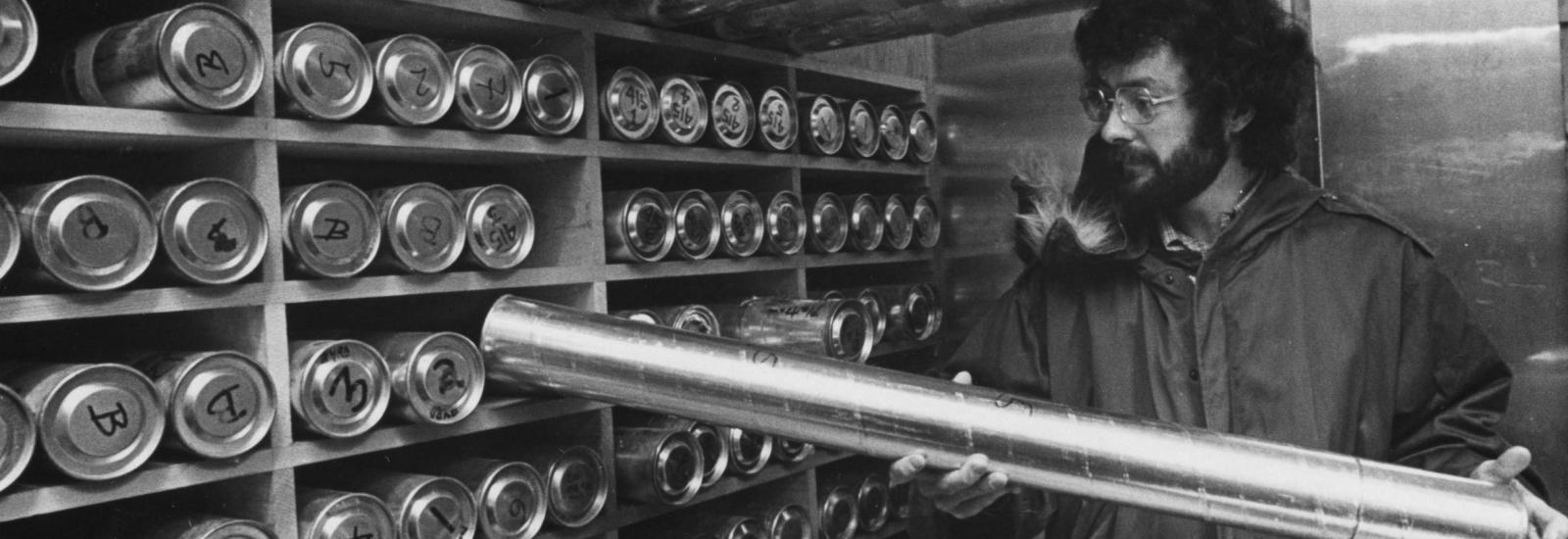 A black and white image of a bearded man with glasses in a winter coat holding a silver cylinder, against a shelf filled with cylinders along the wall.