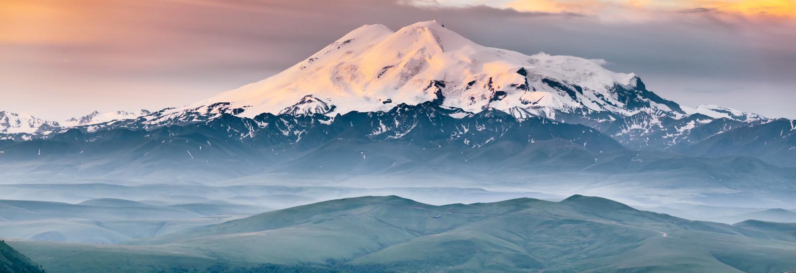 view of Elbrus mount in the morning at sunrise with a dramatic cloudy sky and foggy green highland meadows. Summer mountain landscape. North Caucasus, Karachay-Cherkess Republic, Russia