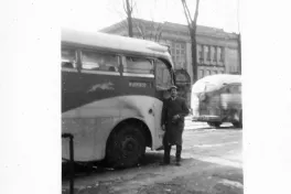 A vintage greyhound bus with a man in a coat standing outside its door. The bus is in a parking lot with a building in the background.